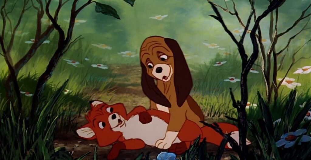 Mały lis Tod i szczeniak Miedziak bawią się razem w trawie – obraz niewinności i braku uprzedzeń.